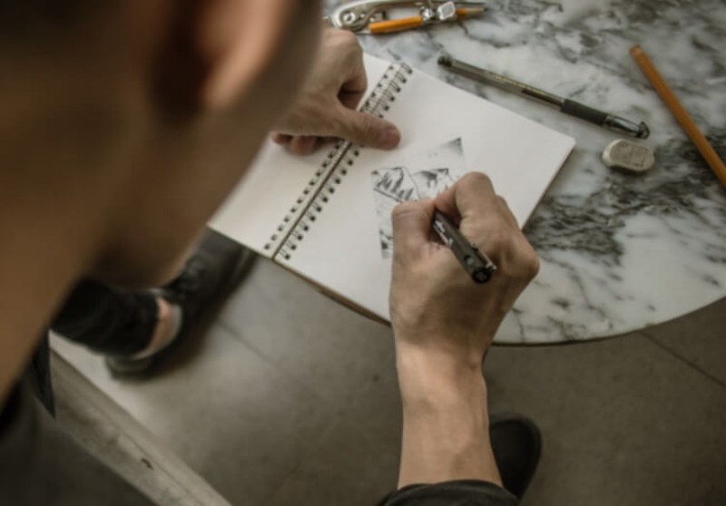 A person is sketching in a notebook on a marble surface, with pencils and a ruler nearby.