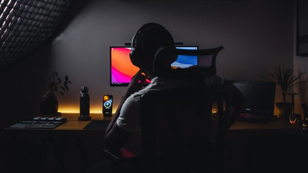 A person with headphones facing a dual-monitor computer setup in a dimly lit room.