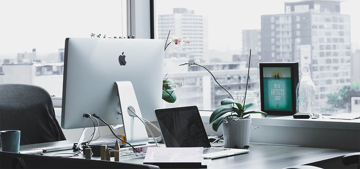 A modern workspace with an Apple iMac, laptop, tablet, plants, and various office supplies set against a backdrop of city windows.