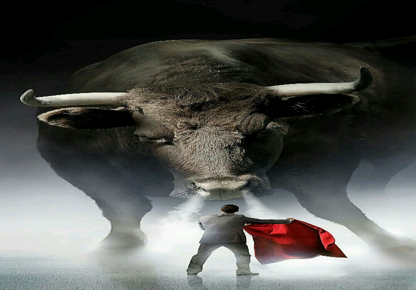 A person in a suit holds a red cloth in front of a large bull amidst a misty backdrop.